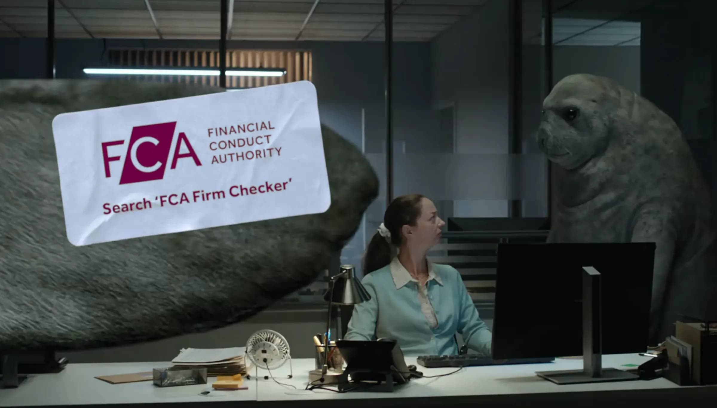 A still from the FCA’s advert: a customer at a desk holding a sign reading “Search FCA Firm Checker”, watched by a seal
